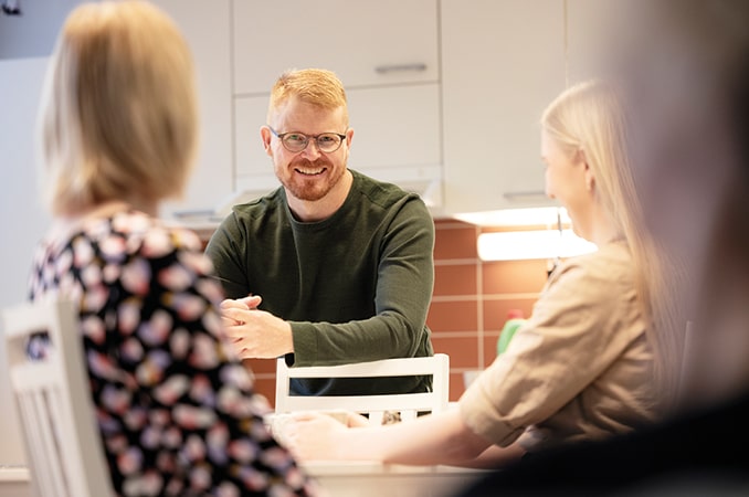 Mies ja kaksi nuorta naista keskustelevat keittiön pöydän ääressä ja mies hymyilee kohti kameraa.