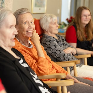 Kolme ikääntynyttä naista istuvat päivähuoneessa. Yksi naisista katsoo kameraan ja hymyilee.